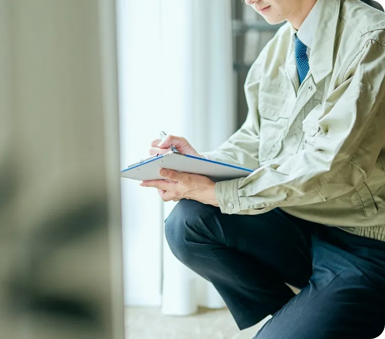 作業服を着た男性がクリップボードにメモを取りながら住宅の点検を行っている様子。