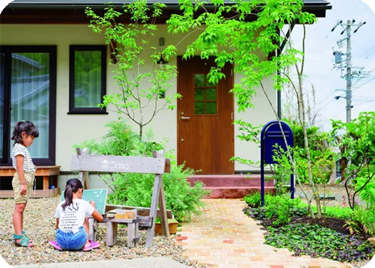 緑に囲まれたナチュラルな庭のある住宅の玄関前で、子どもたちが遊ぶ様子。木製の看板や小さな遊び場が設置され、自然と調和した温かみのある雰囲気。