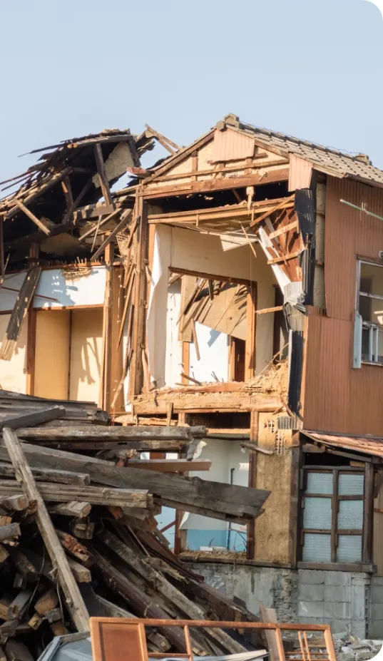 老朽化した建物の解体現場。崩れかけた木造住宅の一部がむき出しになり、手前には解体された木材の残骸が積み重なっている。